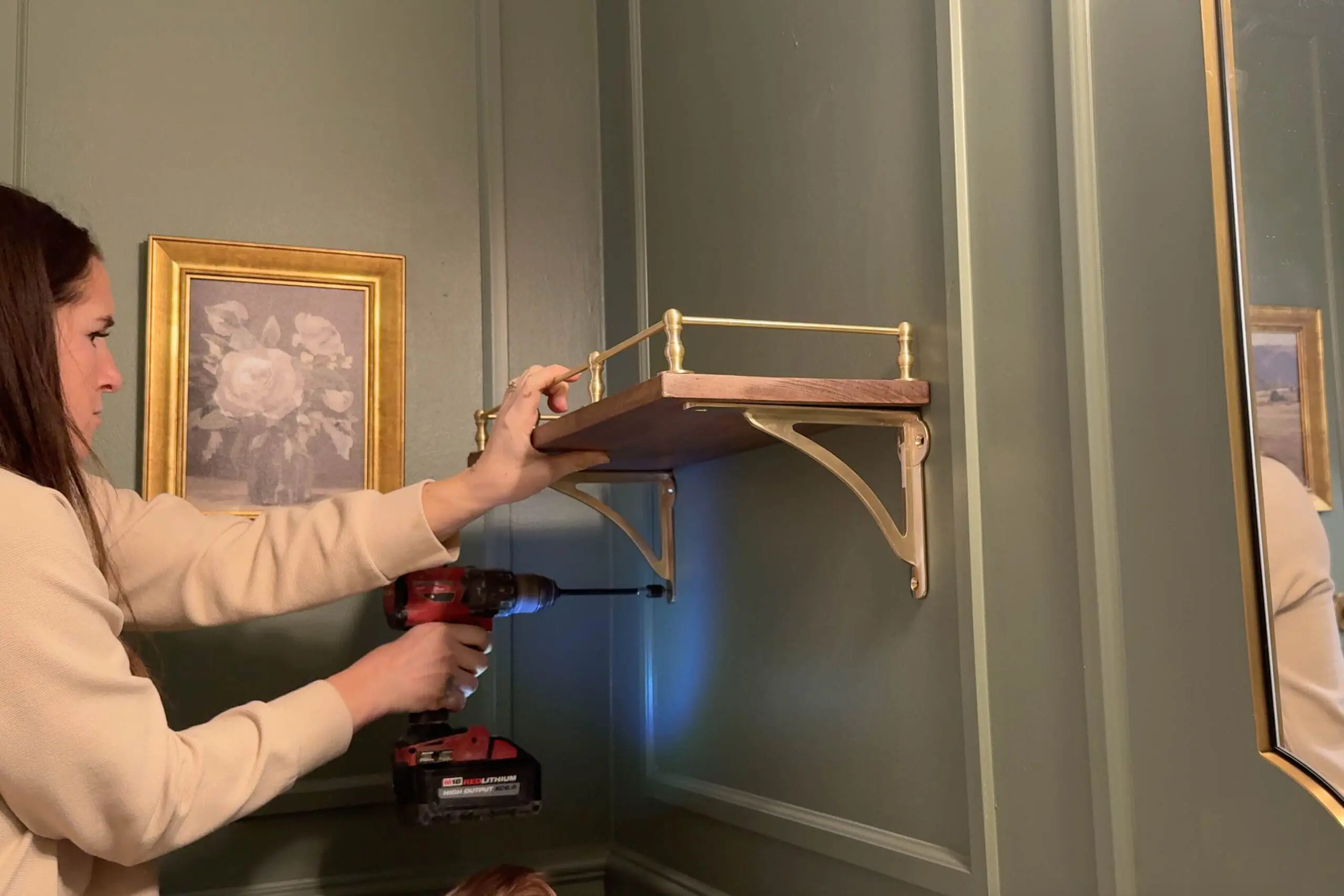 Woman in a cream sweater using a red cordless drill to install a decorative wooden shelf with brass brackets on a dark green bathroom wall, with a framed floral painting visible in the background.